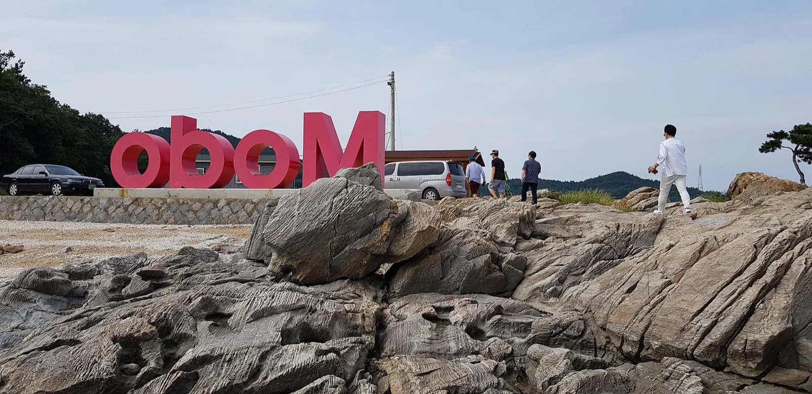 Large red letters spelling Modo standing on a rocky shore, with people walking among the rocks behind them