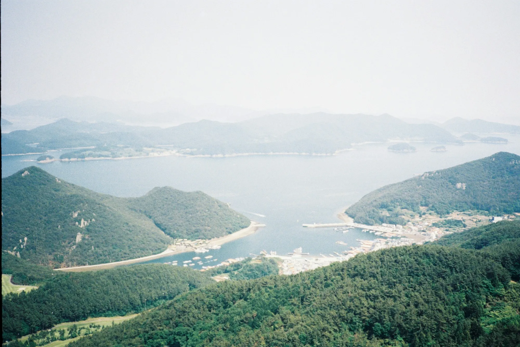 A curved harbour bay with a village, a breakwater and small boats, and forested islands behind